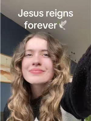 A young woman with long, wavy blonde hair smiles at the camera. Above her, text overlaid reads 'Jesus reigns forever,' accompanied by a small image of a dove. The background shows a view of a room with a blue wall and a window.