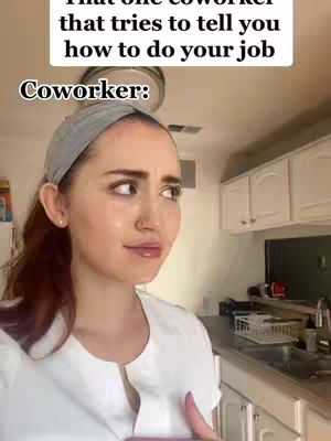 A woman with fair skin stands in a kitchen, looking off to the side with a slightly annoyed expression. The image is a selfie-style shot. The background includes white kitchen cabinets and the edge of a countertop. Above the woman, superimposed white text reads, 'That one coworker that tries to tell you how to do your job'. Below the text at the bottom, in bold is the word 'Coworker:'