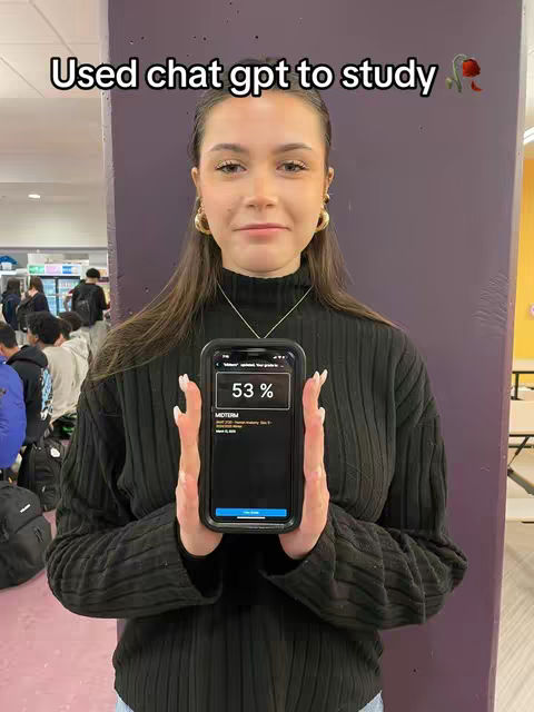 A young woman stands in front of a purple wall, holding a phone that displays a study-related app screen with a 53% grade. Text above reads "Used chat gpt to study". Her hair is down, and she wears a black sweater and large gold earrings. A group of people can be seen in the background. Highlighting the grade.