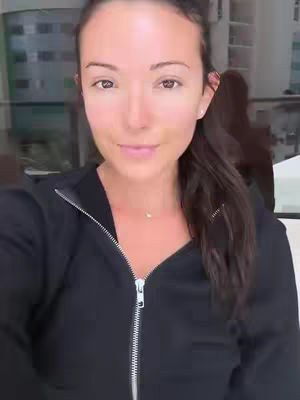A woman with dark hair in a black zip-up jacket smiles at the camera, with a blurred outdoor background.
