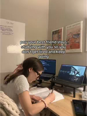 A young woman wearing glasses sits at a desk in a room, studying. Two laptops are open, one with a timer on the display. A book is open, and she is writing. The scene suggests late-night study session.