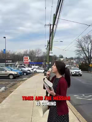 A woman with dark hair in a ponytail, wearing a dark polka-dot dress and a maroon cardigan, stands on a sidewalk speaking into a microphone, holding a Bible. The background includes a suburban street with parked cars, businesses, and cloudy sky, with hook text in red on the bottom of the image.