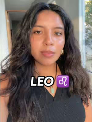 A woman with long, dark, wavy hair is centered in the frame. She gazes towards the camera with a neutral expression. The background is slightly blurred, suggesting an indoor setting. Overlaid on the image are the words 'LEO' in a bold white font, with a purple Leo zodiac symbol next to it. She wears a black top and a necklace. The color palette is natural, with the woman's skin tones and the contrasting black and white text creating visual interest.