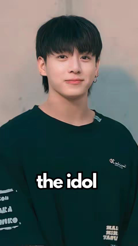 A young man with black hair and a black crew neck sweater is centered in the image, his gaze directed at the viewer. A white text overlay reads "the idol". The background appears to be a plain, light color.