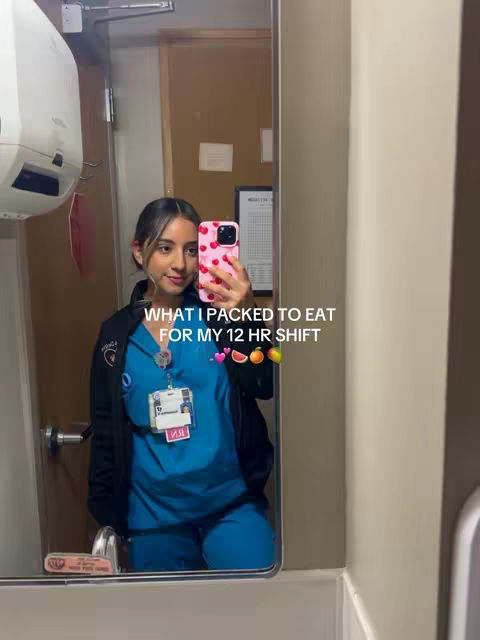 A mirror selfie of a woman in a hospital setting, wearing blue scrubs and a black jacket. The phone she's holding displays a pink case with white polka dots, and the image is framed to include a reflection in the mirror.