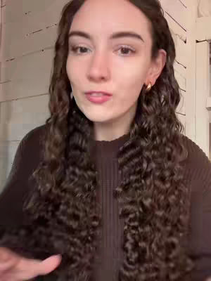 A medium shot features a young person with long, curly brown hair, framed against a light-colored wall. The person looks towards the camera, appearing to be mid-sentence, with a neutral expression. They are wearing a dark brown sweater and small gold hoop earrings. The lighting is neutral, highlighting the person's face and hair.