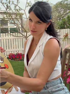 A woman is outside, sitting in front of a white picket fence, with trees and flowers in the backdrop. She's facing the camera, looking slightly to the side with a contemplative expression. She is wearing a white vest over a dark top and is holding a paintbrush over a drawing or painting that is out of the frame. Her dark hair is pulled back.