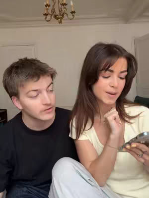 A couple is seated indoors, with a man on the left looking at the woman on the right, who is holding a phone. The setting includes a chandelier and a wall.
