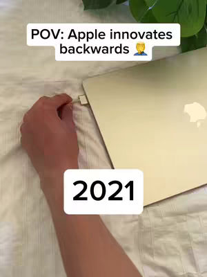 The cover image shows a person's hand connecting a MagSafe cable to a MacBook Pro. Above the laptop is a text overlay that reads 'POV: Apple innovates backwards' along with a person emoji. Below the laptop is a white rectangle with the text '2021'. The scene is on a bed with a white sheet and a green plant peeking from the top right corner.