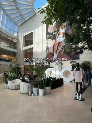 A brightly lit, wide-angle shot capturing a modern, open-plan interior space. The floor is tiled in a light, neutral color, and the architecture features high ceilings with a unique, geometric skylight. A large mural is visible on the wall, depicting a woman and a car. In the foreground, there's an eye-catching display; a mannequin dressed in a light pink outfit. A variety of green plants in white pots are evenly spaced around a room with a sofa and some chairs. The overall atmosphere is modern and upscale.