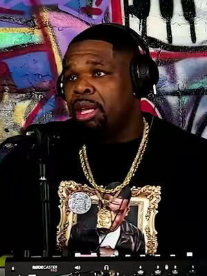 A man with a short, dark haircut and beard is facing forward in front of a colorful graffiti wall. He's wearing headphones and a black t-shirt with a gold chain. The t-shirt has an image on it. A mixing board or audio-related device is visible at the bottom.