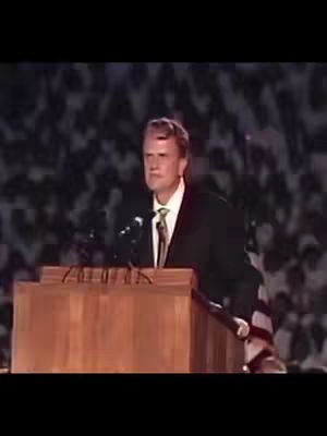 A man in a dark suit, white shirt, and tie stands behind a wooden podium in front of a blurry background of an audience. The man is speaking into a microphone, and the lighting appears to be from overhead.