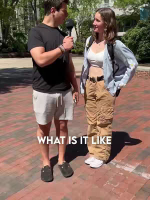 A man wearing a black t-shirt and shorts interviews a woman wearing a white top, cardigan, and khaki pants. The image is taken outdoors, possibly on a college campus, with text overlayed at the bottom.