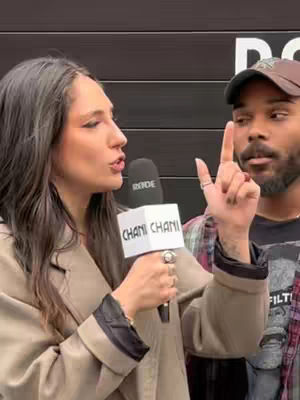 A woman with long, graying hair is holding a microphone and speaking, facing left. A Black man wearing a cap is on the right looking at her, pointing one finger up. The backdrop shows a dark, textured surface.