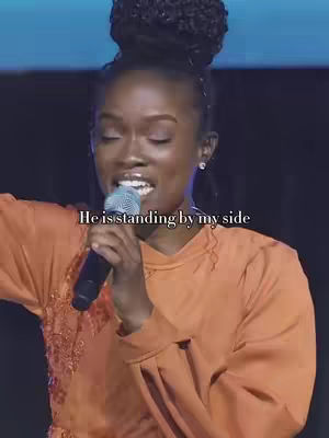 A medium shot focuses on a Black woman singing into a microphone. She wears an orange top and has her hair styled in a high bun. The text overlay reads, "He is standing by my side."