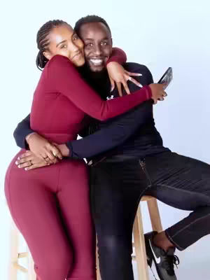 A candid photo of a couple embracing, the woman in a maroon top hugging the man who is holding a phone. They are on a white background with a wooden stool.