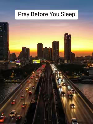 The image shows a cityscape at dusk or dawn. The sky transitions from orange near the horizon to a darker blue. There are several tall buildings silhouetted against the colorful sky. A bridge, with multiple lanes of traffic, dominates the foreground. The cars are moving with their headlights on. The overall effect is one of urban life, illuminated by the setting sun.