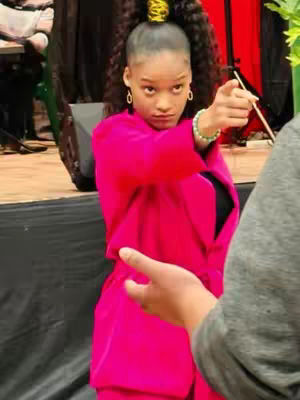 A woman in a pink suit gestures emphatically with a serious expression, as if making a pronouncement. A hand is in the foreground, partially obscuring the view. The background is dark and out of focus, but there are hints of a stage or event setting.