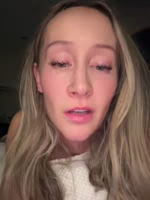 A close-up shot of a woman, with her face dominating the frame. She has long, wavy blonde hair, and her expression appears concerned or emotional. Her eyes are looking downward, and her lips are slightly parted. The background is dark, suggesting a low-lit setting.
