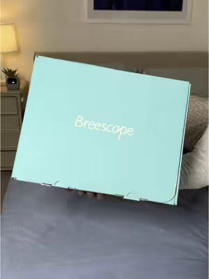 A box is being held in front of a gray bed. The box is light blue with the word "Breescope" in a handwritten-style font. The background includes a bedside table with a lamp and a window.