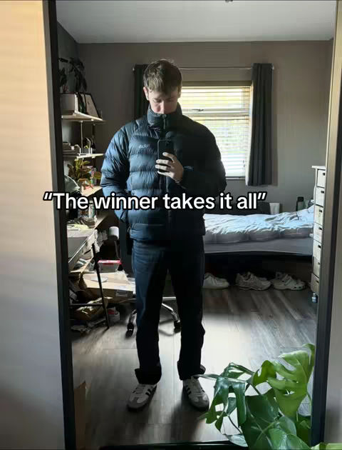 A full-length mirror shot captures a man standing in a room reflecting his image. He appears to be taking a selfie. Behind him are various elements of a bedroom, including the bed and a window with blinds. The room is lit with natural light. The man is dressed in dark-colored clothing, creating contrast with the lighter tones of the room. A plant is visible in the foreground, adding a natural element to the scene.