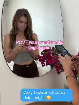 A woman stands in front of a rounded mirror, looking at her phone. Text overlays appear on the image. In the foreground, the woman is holding a phone and there are flowers. A speech bubble is visible.