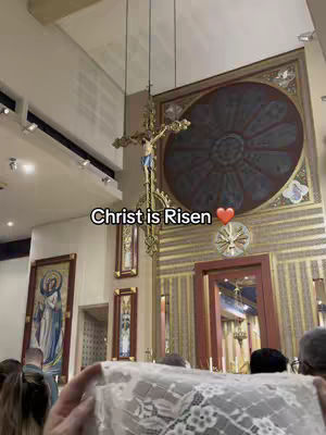 The cover image shows a view inside a church during a religious service. In the foreground, a person holds a white lace cloth. The background features a gold crucifix hanging from the ceiling and a large, ornate wall painting. The text overlay says Christ is Risen.