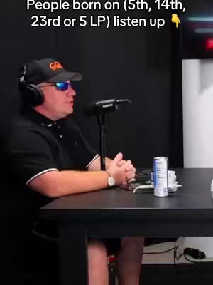 A man in a black shirt and a baseball cap sits at a table with a microphone in front of him. He is wearing sunglasses and there is text overlayed on the top of the image.