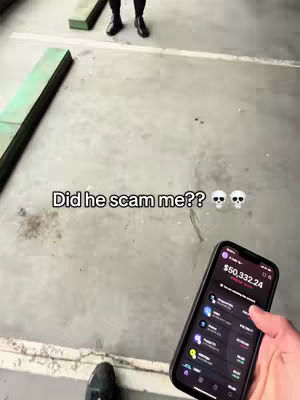 The cover image is a vertical shot taken from a low angle. It shows a person's hand holding a phone displaying what appears to be a cryptocurrency trading app. The phone screen shows the amount of $50,332.24, along with various cryptocurrency icons and financial data. In the background, out of focus, are the legs and feet of a person standing on a concrete surface next to green-colored beams. Above the phone screen and centered in the image are the words "Did he scam me??" along with two skull emojis. The overall tone seems to be a mix of shock and financial concern, given the displayed text and emojis.