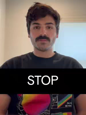 A medium shot features a man with dark hair and a mustache, positioned in front of a neutral background. The man is wearing a black t-shirt, and a black rectangle covers the lower portion of his torso with white text overlayed on it. The background appears to be a light-colored wall and door.