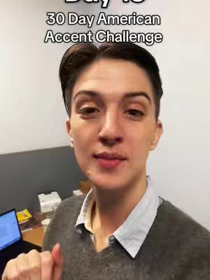 A woman with short dark hair and a grey sweater is centered in the frame, in front of a light-colored wall. The text overlay at the top reads "Day 13 30 Day American Accent Challenge."