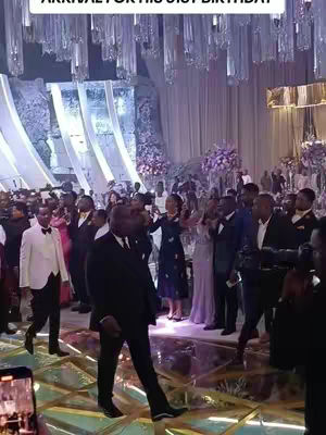 A wide shot captures a grand, decorated ballroom setting, with soft lighting and numerous people gathered. A man in a black suit walks forward, and a crowd of people in formal attire are visible, with decorations hanging from the ceiling.