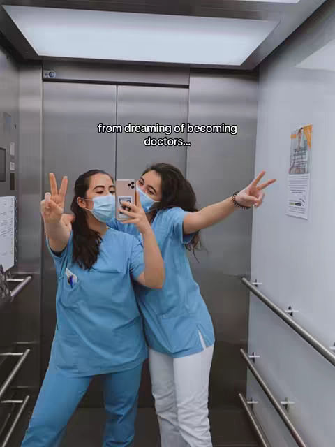 Two young women in medical scrubs pose for a selfie inside an elevator. One woman holds the phone and the other raises a peace sign. The background is a metallic elevator.