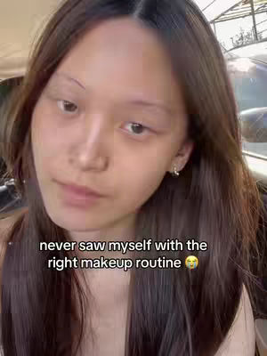 A selfie-style video featuring a young woman with long, dark brown hair. She is looking at the camera with a slight tilt of her head. Overlay text at the bottom reads: 'never saw myself with the right makeup routine' with a crying face emoji. The background appears to be a somewhat blurred outdoor setting.