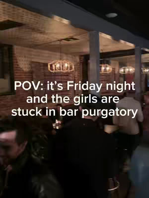 A vertical video shot inside a bar. The scene is bustling with people, though details are somewhat obscured. The foreground shows blurry figures moving around; a portion of someone's face is visible. In the midground, a bar setup with drinks. The background reveals the interior design, featuring exposed brick walls and ornate, warm-toned chandeliers. Soft lighting creates a slightly dim, ambient atmosphere.