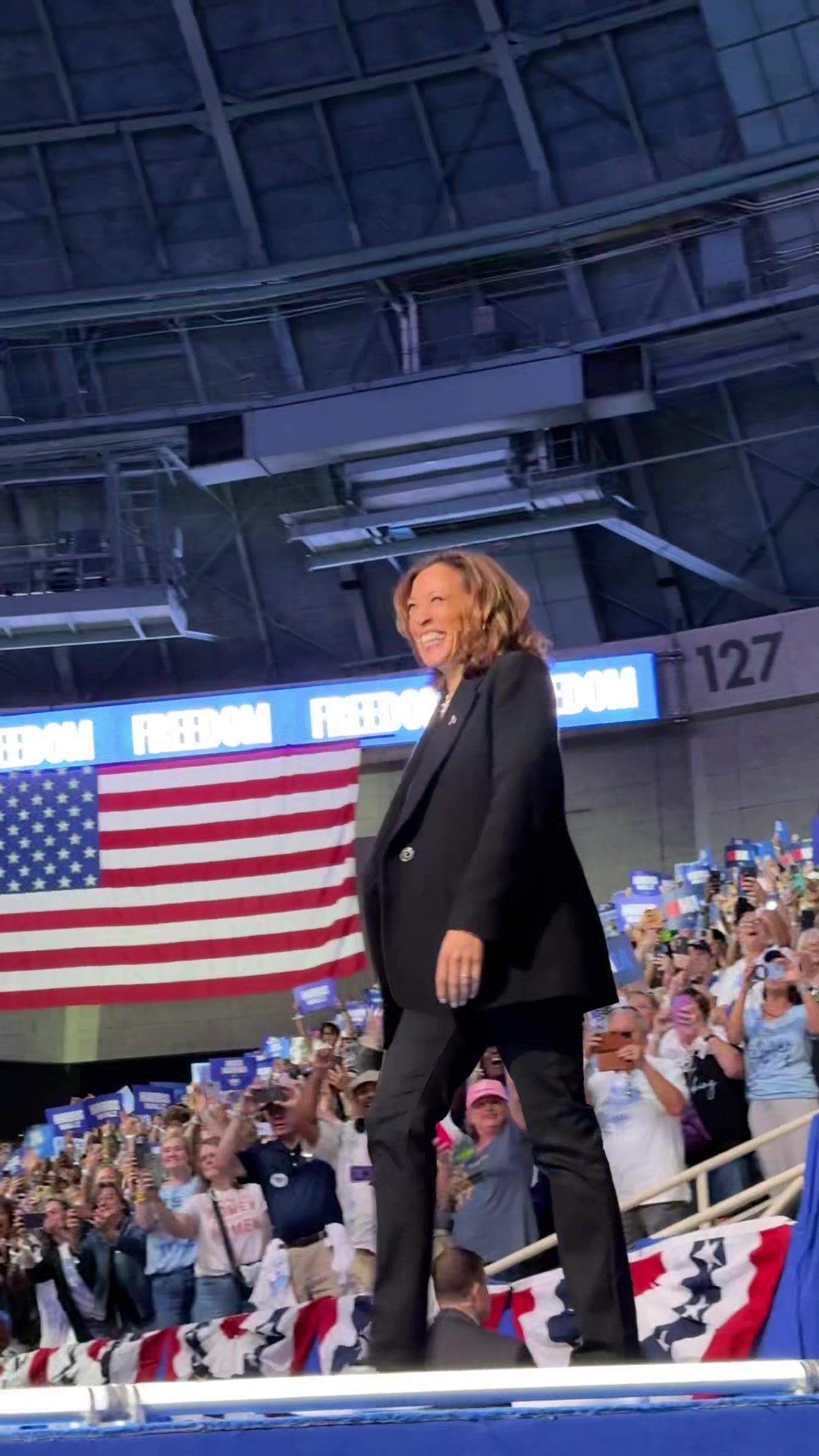 The cover image features a medium shot of Kamala Harris walking on a stage in front of a large American flag. She is smiling and looking off to the side. Behind her is a crowd of people, many holding up signs. The background includes a banner with text and overhead lighting.