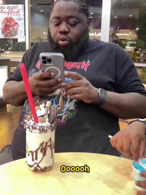 A man holding a phone and looking at it with a surprised expression, a large milkshake in foreground.
