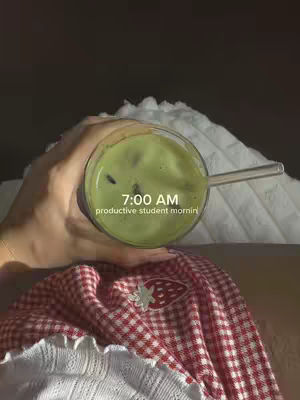 A hand holds a glass of green drink, possibly matcha or a similar beverage, against a backdrop of a white, textured surface, likely a bed or blanket. A red and white checkered fabric, perhaps a blanket or a shirt, is visible, and the overall composition is warm and inviting.