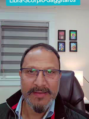 A man with glasses and a beard is centered in the frame, with a background containing blinds, a window, and framed pictures.