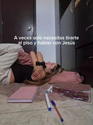 A Young woman is lying on the floor, her head resting on a pillow, and holding another pillow close to her. A notebook, pens, and papers are spread around her. The scene suggests introspection and possibly prayer.