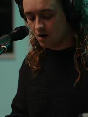 The image is a medium shot focusing on a person with long, curly brown hair and wearing a black top. The person is positioned slightly off-center to the right, with a microphone in front of them on the left. They have headphones on, and their eyes are closed. They appear to be singing or speaking, with their mouth open and directed towards the microphone. The background is blurred with a light teal color.