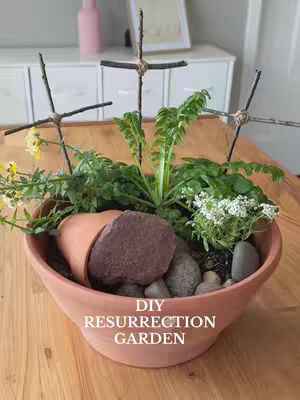 A terracotta bowl contains a DIY resurrection garden. The garden features a small terra cotta pot on its side, a large brown rock, and medium-sized pebbles to represent the tomb. Three crosses made of twigs are placed throughout the garden along with green plants. The text 'DIY RESURRECTION GARDEN' is written in white text at the bottom. The background shows a wooden table and a white cabinet.