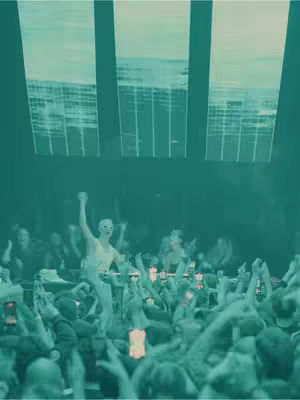 A concert scene bathed in green tones, showing a person on stage in a white outfit, possibly a performer, with their arm raised. A large crowd of people is visible, many holding up phones. The background features large, illuminated panels.