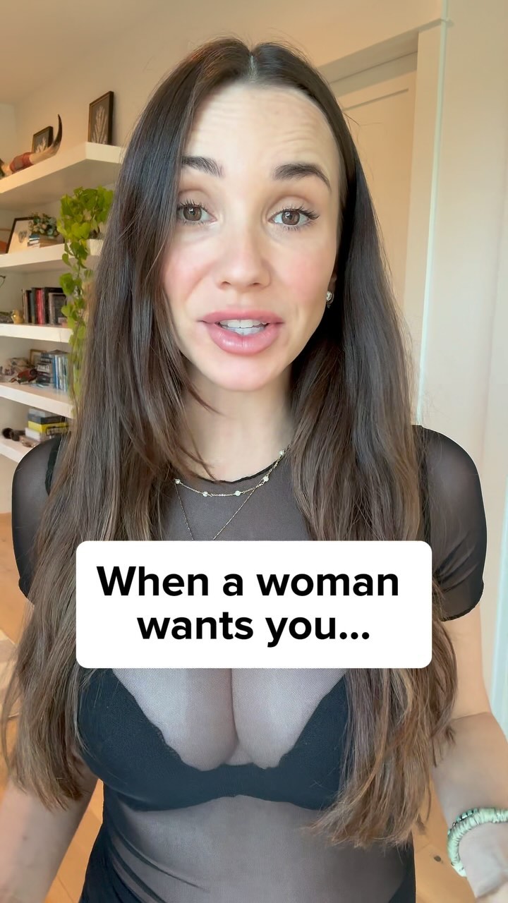 A woman with long brown hair and makeup is centered in the frame, looking at the camera and talking. A white rectangle with black text overlay is prominently featured in the lower third. The background includes a glimpse of a room with shelves and natural lighting, giving the impression of a home setting.