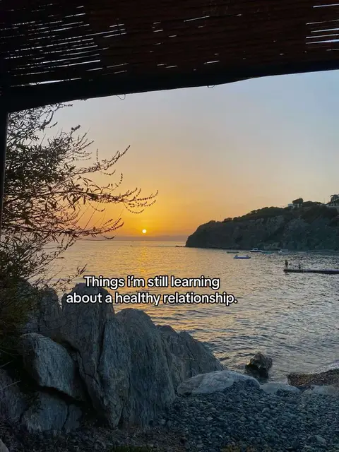 The image captures a serene sunset over a tranquil bay. The sky is a gradient of orange and blue, with the sun low on the horizon. In the foreground, there are rocks and a body of water, and the edges of a structure can be seen at the top of the frame. The overall feeling is one of peace and natural beauty.