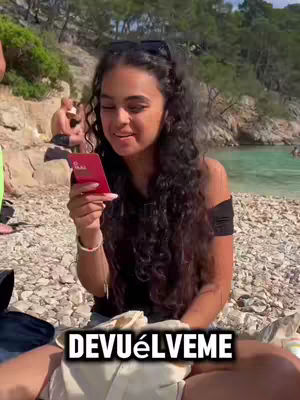 A woman with long dark hair sits on a sandy beach, holding a red card in her hands. The background includes the ocean, other people, and some rocks. A text overlay at the bottom reads 'DEVUÉLVEME'.