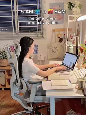 A woman with long black hair sits at a desk in a well-lit room facing a laptop. She is wearing glasses and a white t-shirt. In the background is a bright window.