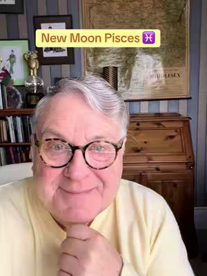 An older man with glasses smiles, angled to the camera. He is wearing a yellow collared shirt. Behind him is a wooden cabinet, a map, framed pictures, and a bookshelf with books.