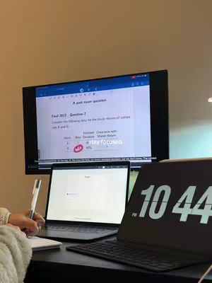 The image shows a desk with study materials. A large monitor displays a document, a laptop is open in front of the monitor. A notepad is close to a pen and a timer.