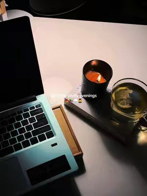 An overhead shot displays a study setup on a white surface. A laptop occupies the left, its screen dark, with the keyboard visible. A black candle with a lit flame and a glass of tea with a lemon slice are to the right. A notebook or book rests between them, with text overlayed on the image 'Autumn study evenings'. The overall composition is warm, lit by the candle, evoking a cozy, autumnal atmosphere.